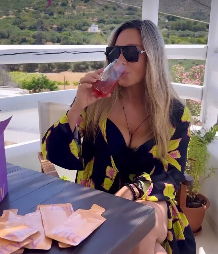 Das Bild zeigt die blonde Fitnessinfluencerin Eleni Petroulaki, sie trägt eine Sonnenbrille und sitzt auf einer Terrasse. Sie trägt ein schwarzes Kleid mit gelbem Blumenmuster. In der Hand hält sie ein Glas mit hajoona Vitality. Auf dem Tisch liegen hajoona Vitality Sachets. Neben den Sachets steht die lila Vitality Produktverpackung. Im Hintergrund sind grüne Hügel zu sehen.