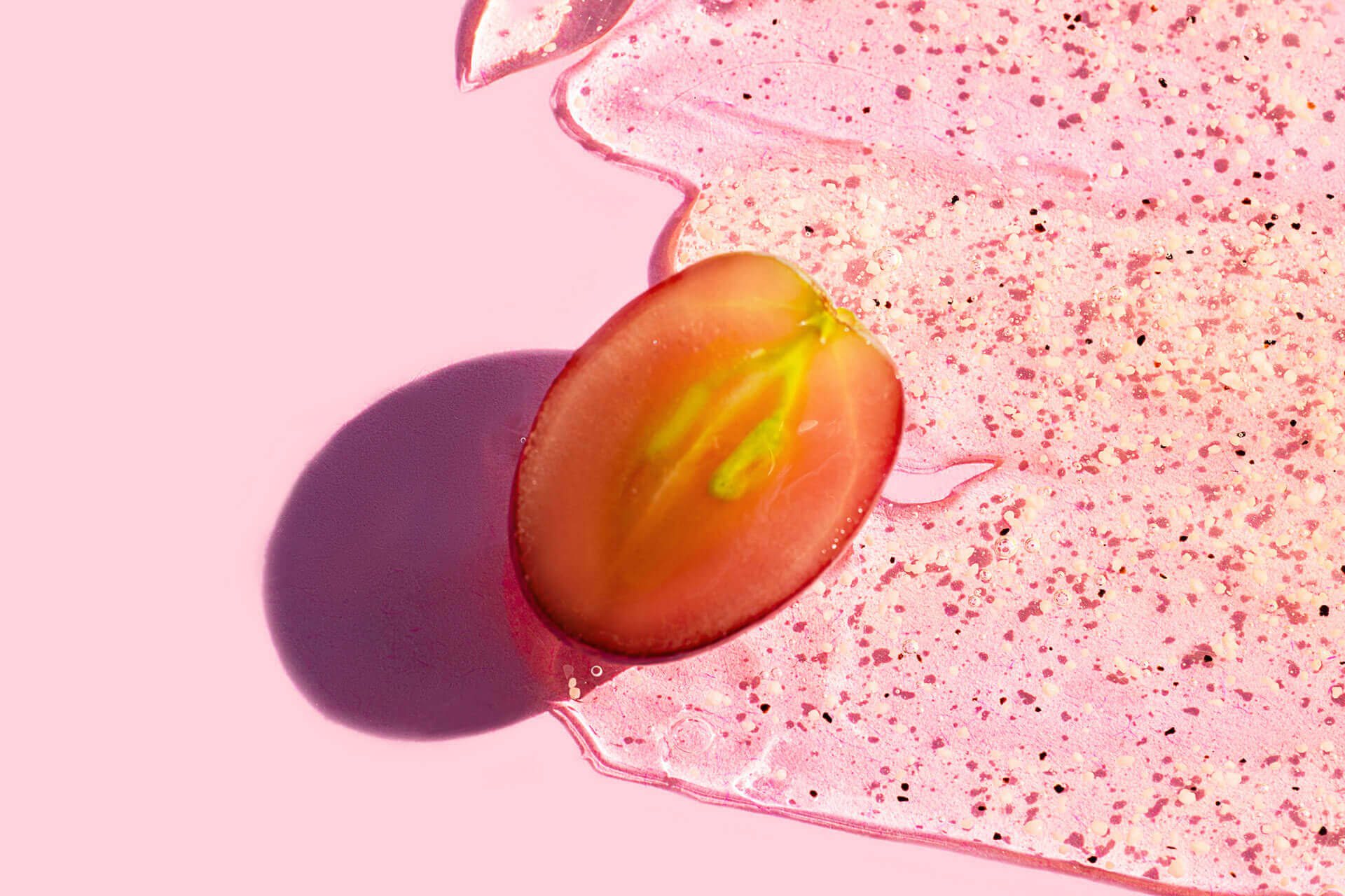 Eine rote Traube, der Länge nach halbiert, liegt auf einer rosa Fläche. Neben ihr ist eine glänzende, rosafarbene Gelmasse mit kleinen dunklen Partikeln zu sehen. Die Szene ist in kräftigen Farben gehalten und vermittelt Frische und Natürlichkeit.